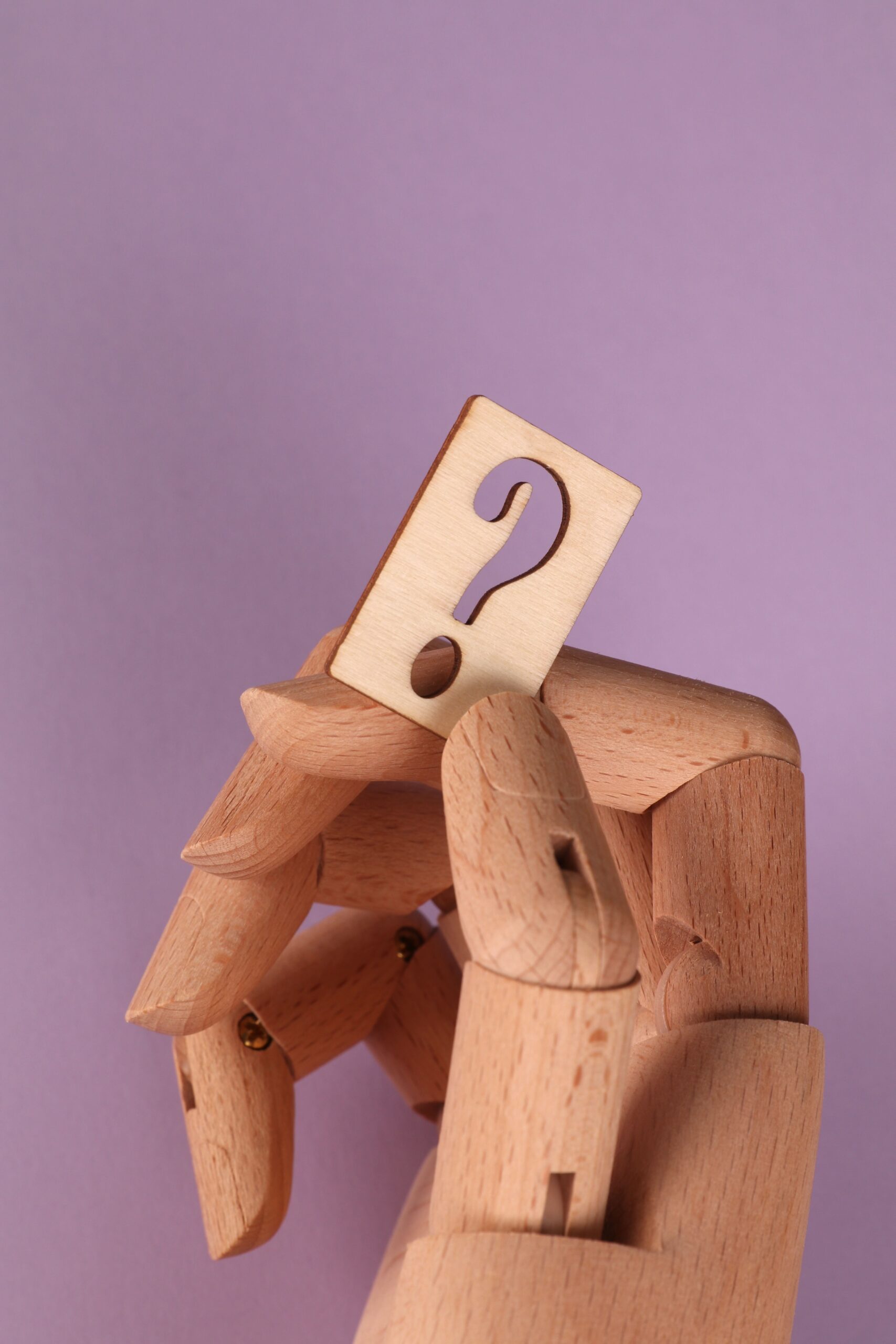 Wooden mannequin hand holding a carved question mark against a purple background.