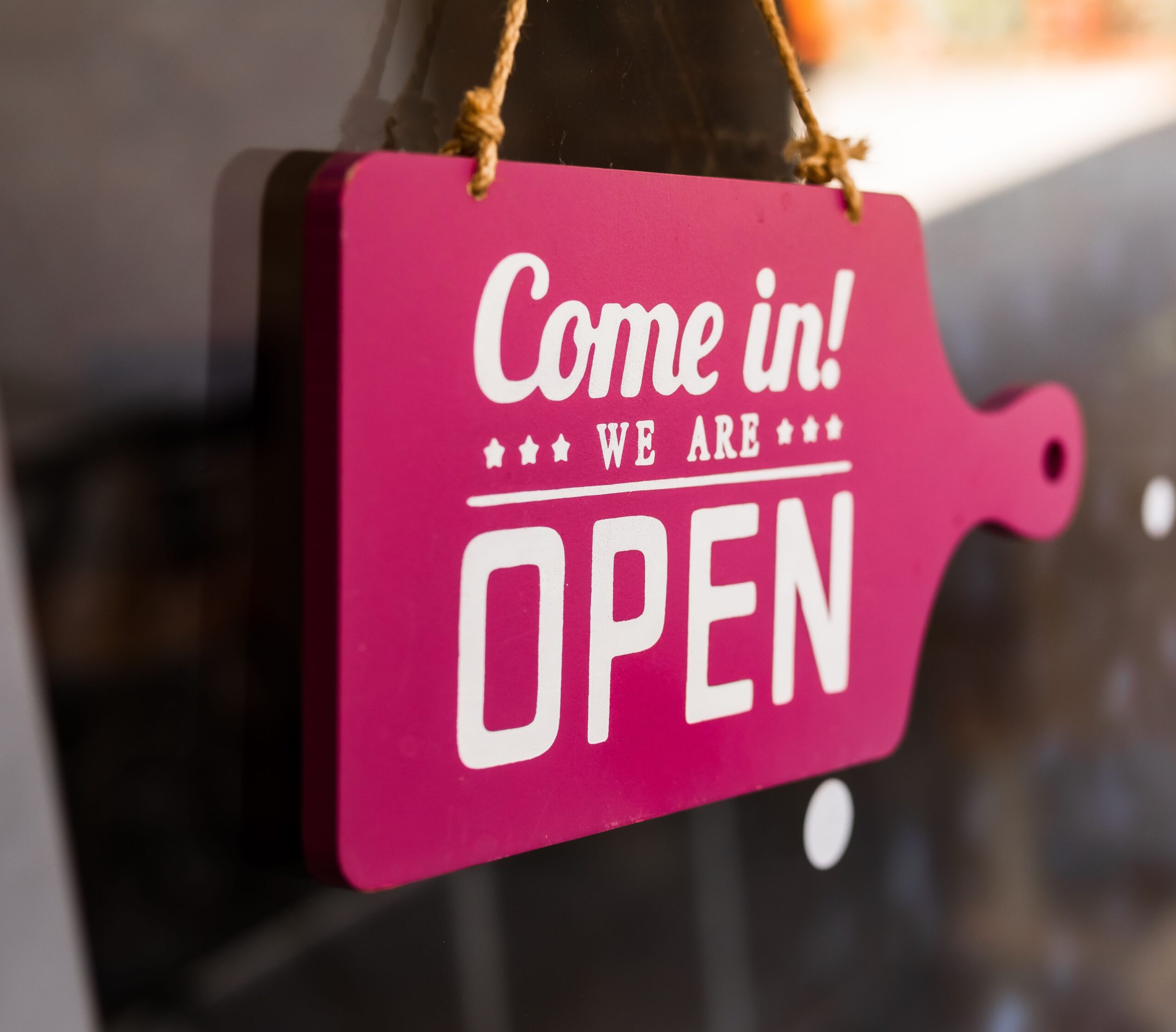 A bright pink "Come in! We are Open" sign hanging in a store window welcoming customers.